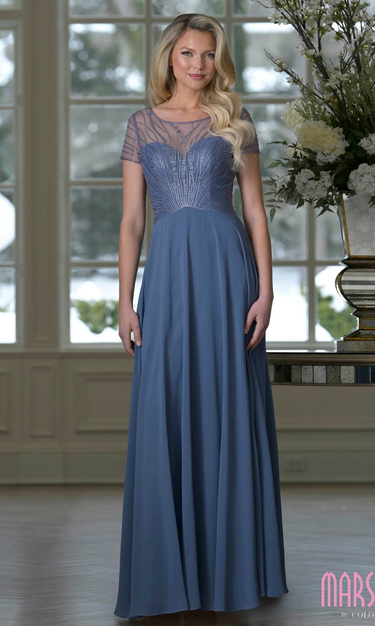 A woman wears the Marsoni M351, a slate blue floor-length chiffon A-line gown with short sleeves and a sheer beaded bodice, perfect as an elegant mother-of-the-bride dress. Large floral arrangements and tall windows are behind her.
